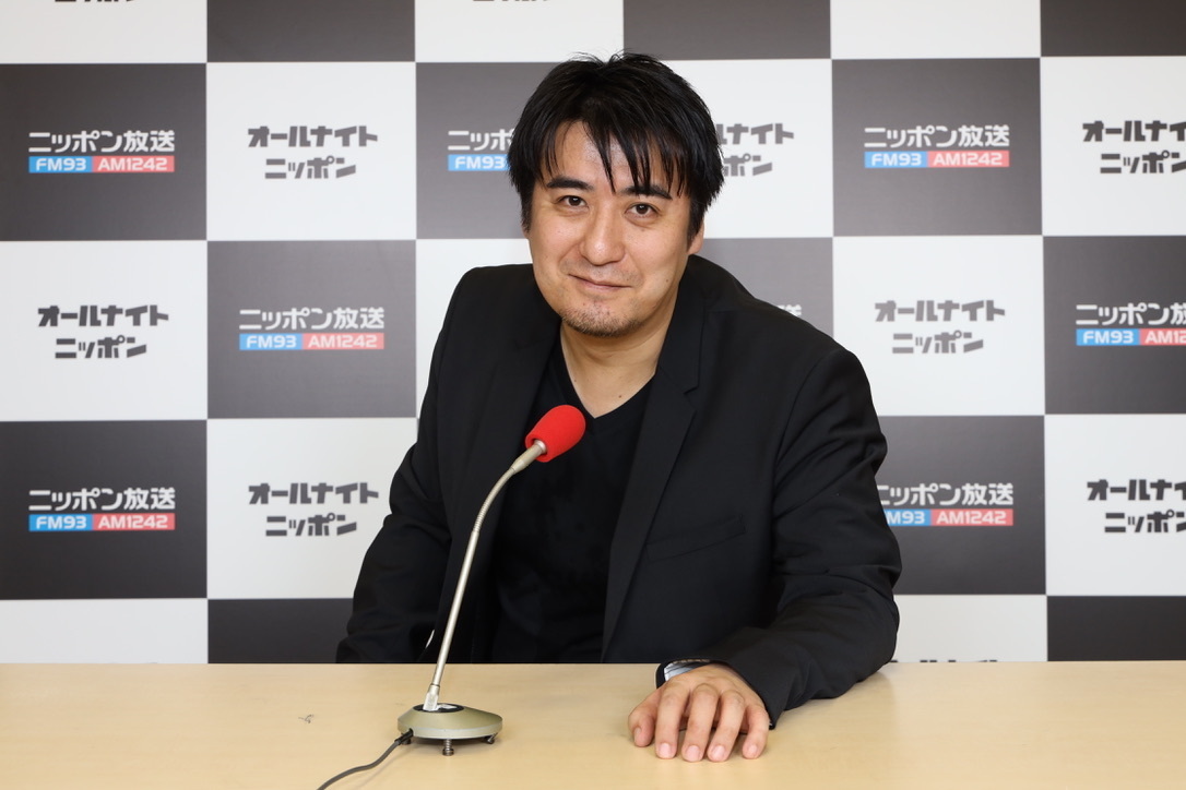 ラジオの宣伝パネルを背景に、マイクの前に座る佐久間宣行さん